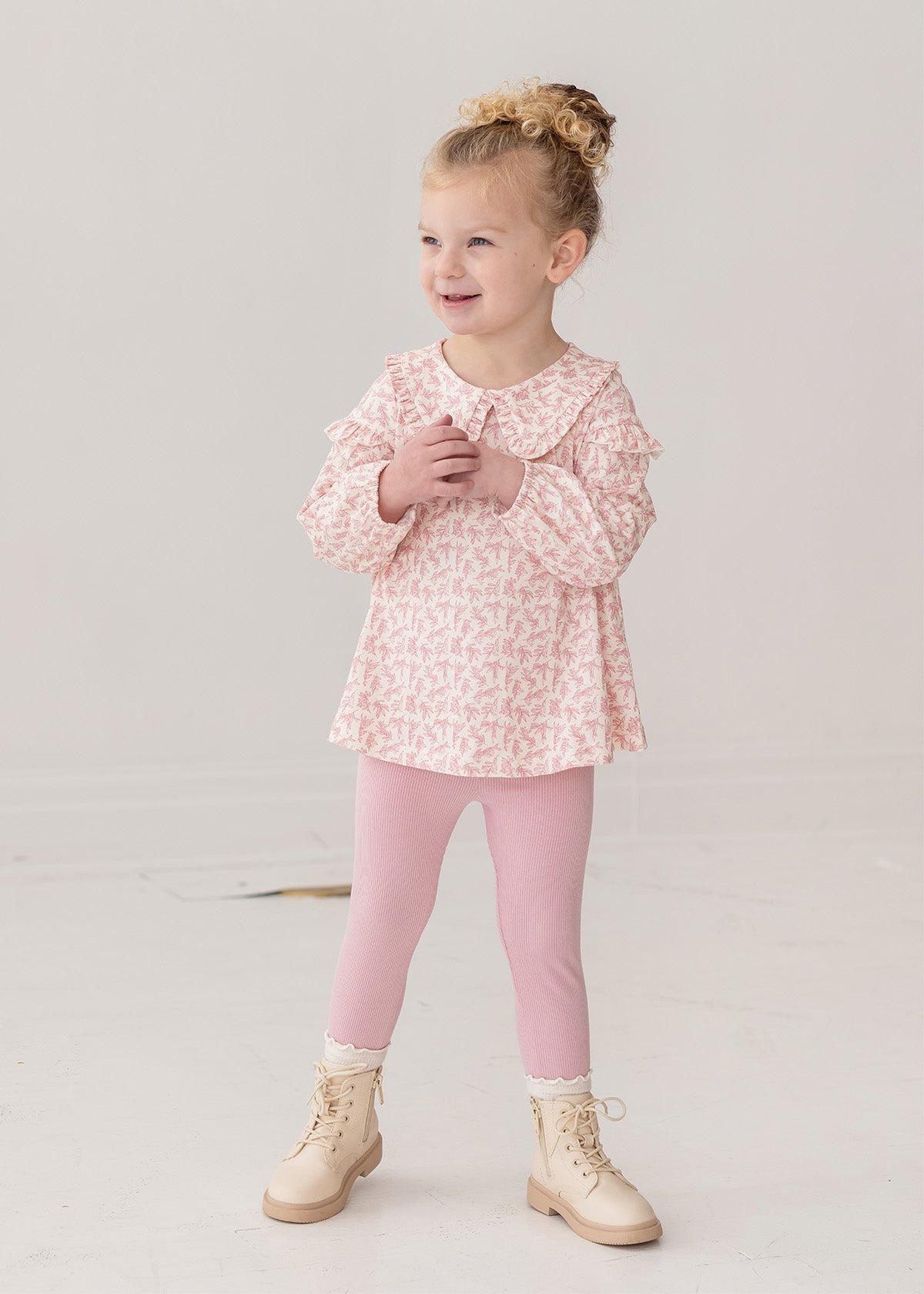 A young girl with curly blonde hair in a bun smiles while wearing the Mabel and Honey Primrose Two Piece Set—featuring a floral top and matching pink leggings—an adorable baby girl outfit, set against a plain white background.