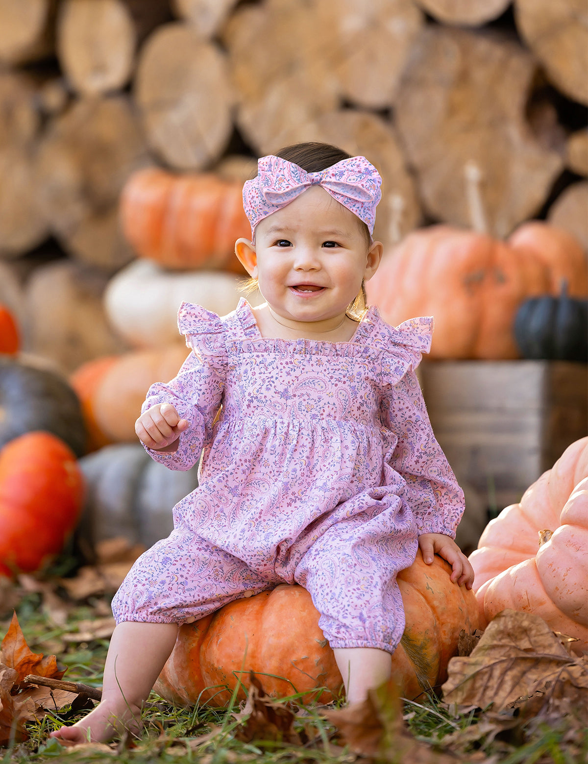 A smiling baby wearing the Mabel and Honey Paisely Romper with a matching headband sits on a pumpkin outdoors, surrounded by autumn leaves, pumpkins, and stacked firewood.