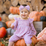 A smiling baby wearing the Mabel and Honey Paisely Romper with a matching headband sits on a pumpkin outdoors, surrounded by autumn leaves, pumpkins, and stacked firewood.
