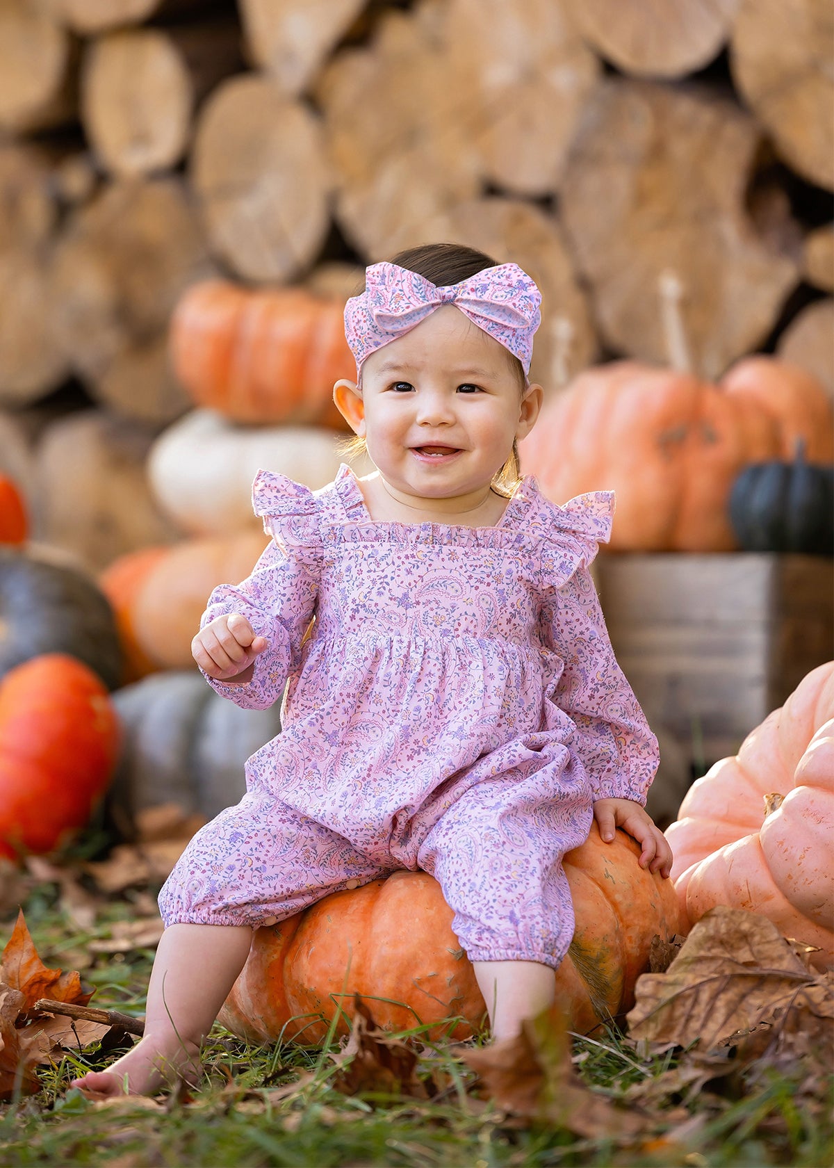 A smiling baby wearing the Mabel and Honey Paisely Romper with a matching headband sits on a pumpkin outdoors, surrounded by autumn leaves, pumpkins, and stacked firewood.