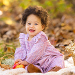 A smiling baby with curly hair wears the Paisely Romper by Mabel and Honey, sitting on a cream blanket outdoors among autumn leaves, holding a red and yellow apple next to a small basket of white flowers.