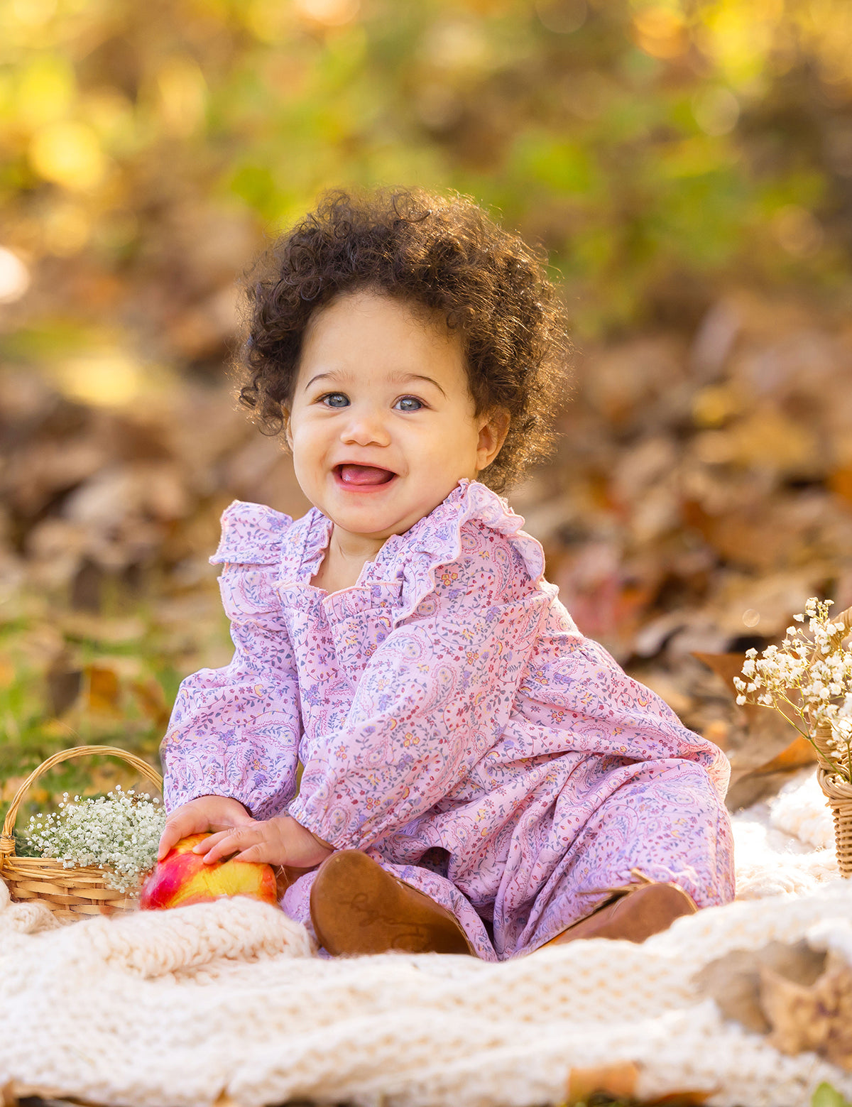 A smiling baby with curly hair wears the Paisely Romper by Mabel and Honey, sitting on a cream blanket outdoors among autumn leaves, holding a red and yellow apple next to a small basket of white flowers.