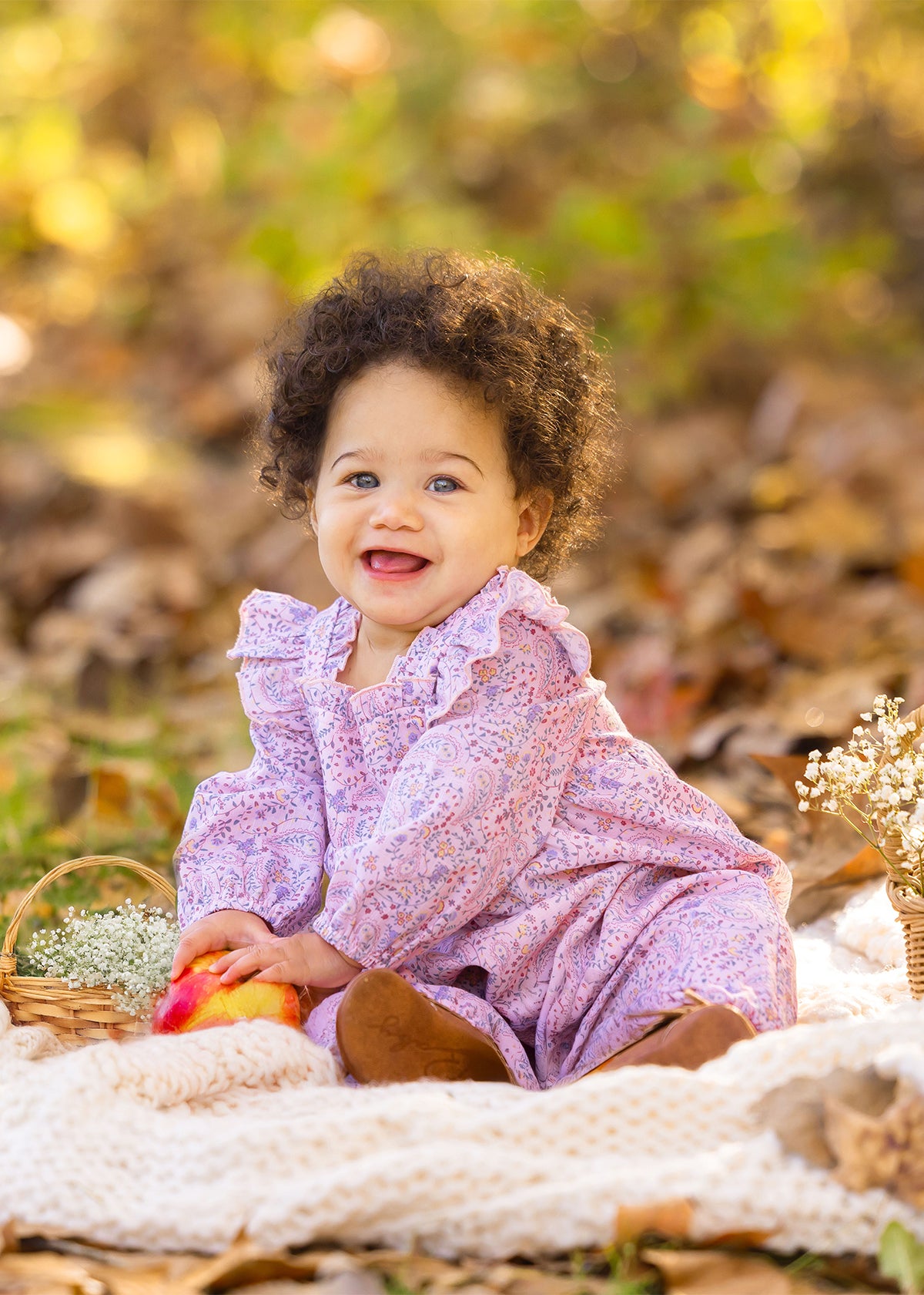 A smiling baby with curly hair wears the Paisely Romper by Mabel and Honey, sitting on a cream blanket outdoors among autumn leaves, holding a red and yellow apple next to a small basket of white flowers.