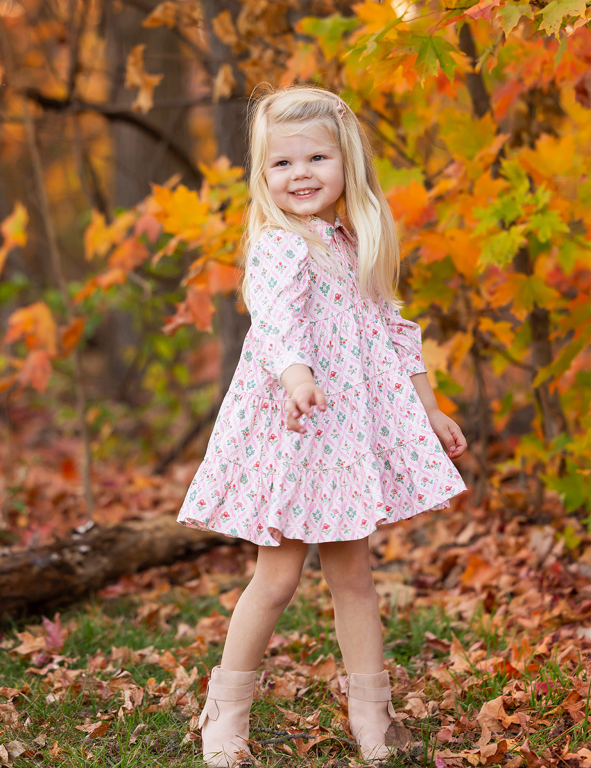 A young girl with long blonde hair smiles outdoors among autumn leaves, wearing the Dixie Pink Floral Dress by Mabel and Honey with a tiered silhouette and light boots.