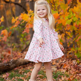 A young girl with long blonde hair smiles outdoors among autumn leaves, wearing the Dixie Pink Floral Dress by Mabel and Honey with a tiered silhouette and light boots.