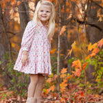 A young girl with long blonde hair, wearing the Mabel and Honey Dixie Pink Floral Dress and tan boots, stands on a log in a forest filled with colorful autumn leaves. She smiles and gazes off to the side.