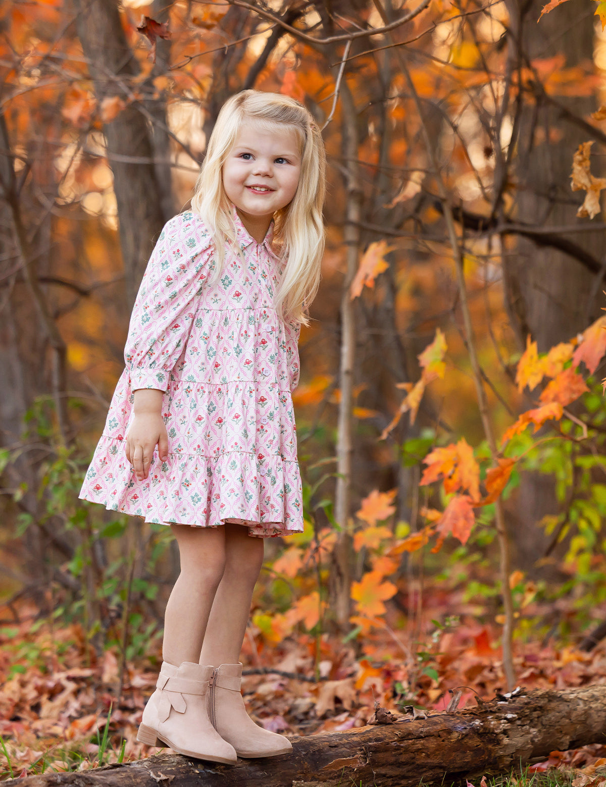 A young girl with long blonde hair, wearing the Mabel and Honey Dixie Pink Floral Dress and tan boots, stands on a log in a forest filled with colorful autumn leaves. She smiles and gazes off to the side.