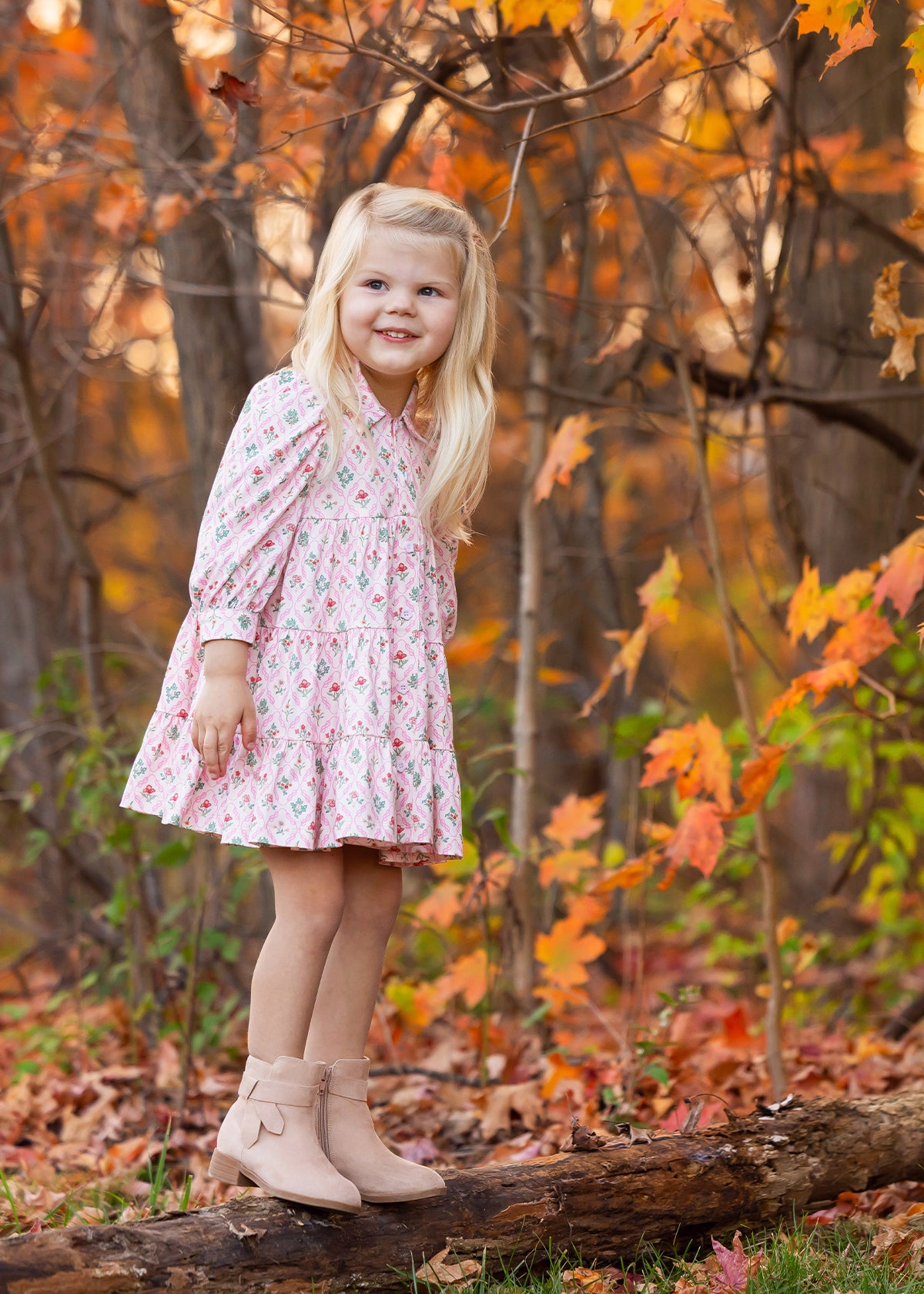 A young girl with long blonde hair, wearing the Mabel and Honey Dixie Pink Floral Dress and tan boots, stands on a log in a forest filled with colorful autumn leaves. She smiles and gazes off to the side.