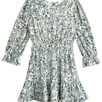 The Brooklyn Floral Dress by Mabel and Honey is a long-sleeve white dress with a green and blue floral pattern, cinched waist, ruffled cuffs, and ruffled hem.