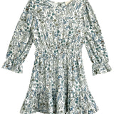 The Brooklyn Floral Dress by Mabel and Honey is a long-sleeve white dress with a green and blue floral pattern, cinched waist, ruffled cuffs, and ruffled hem.