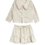 The Juliette Two Piece Set by Mabel and Honey is a charming girls’ outfit with a ruffled-collar button-up top and drawstring skirt, both in lightweight fabric with a delicate floral print.