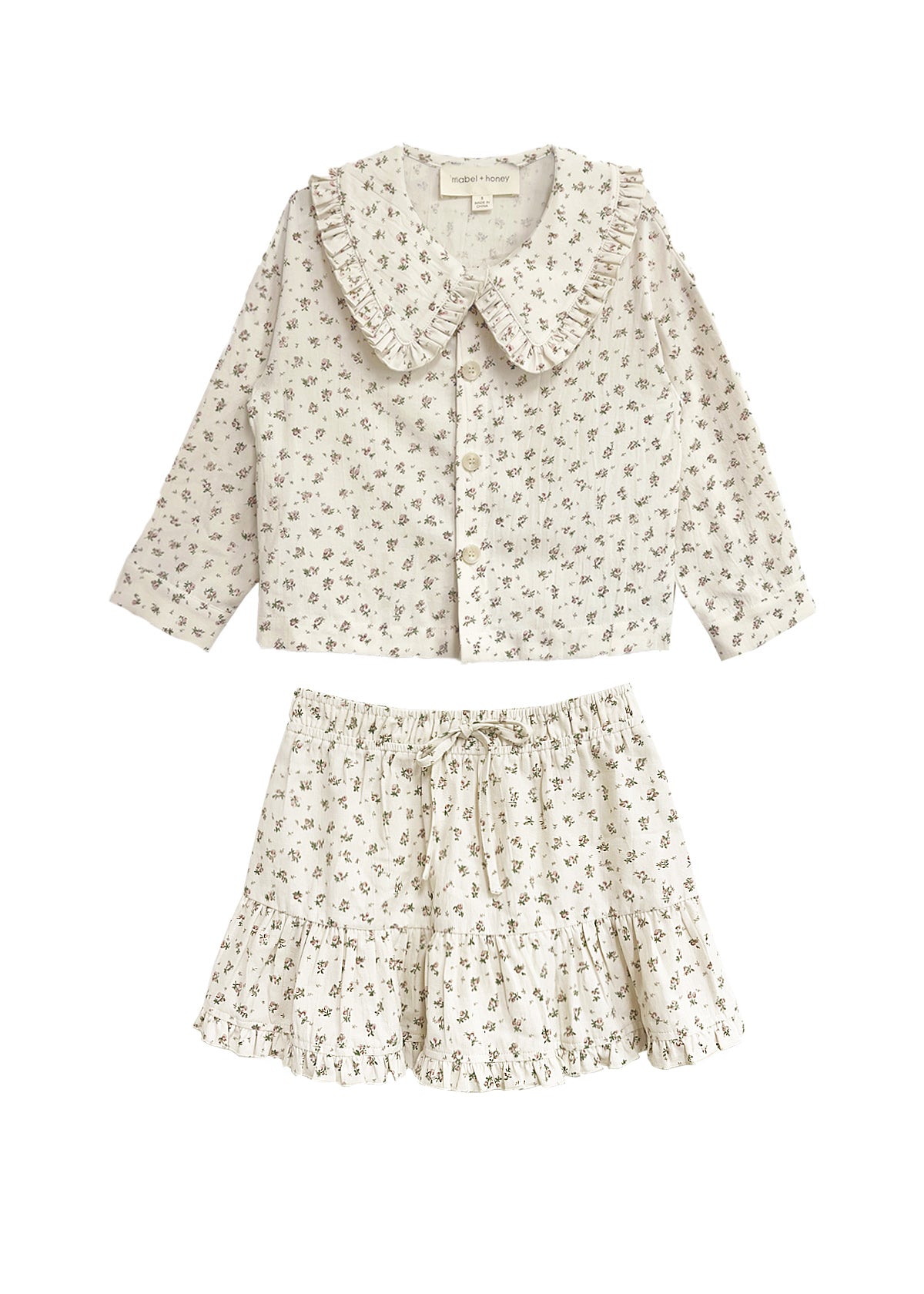 The Juliette Two Piece Set by Mabel and Honey is a charming girls’ outfit with a ruffled-collar button-up top and drawstring skirt, both in lightweight fabric with a delicate floral print.