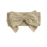 The Cinnamon Sugar Hair Accessory by Mabel and Honey is a beige fabric headband with a large bow and subtle textured floral pattern, adding elegant charm to any outfit.