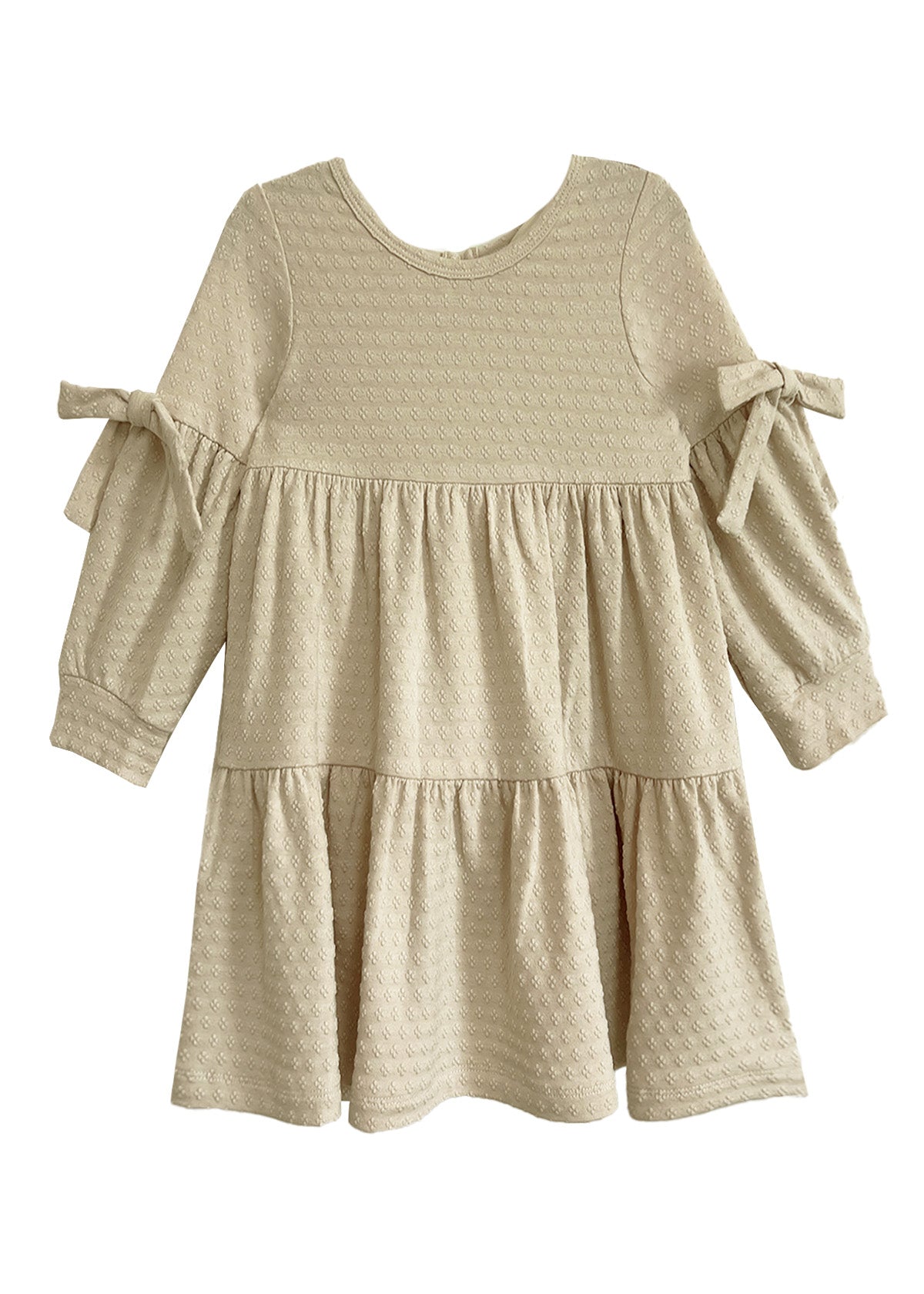 The Mabel and Honey Cinnamon Sugar Dress is a cream-colored toddler dress with textured fabric, a tiered silhouette, long sleeves, and decorative bows on each sleeve.