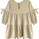 The Cinnamon Sugar Dress by Mabel and Honey features a cream, long-sleeve tiered design with textured dot fabric, ruffled skirt, small bow ties on the arms, and a keyhole button closure at the back for easy dressing.