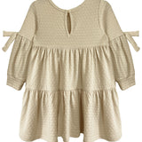 The Cinnamon Sugar Dress by Mabel and Honey features a cream, long-sleeve tiered design with textured dot fabric, ruffled skirt, small bow ties on the arms, and a keyhole button closure at the back for easy dressing.