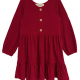 The Mabel and Honey Rosie Dress is a red, long-sleeve girl’s dress with ribbed fabric, wooden front buttons, a round neckline, and a gathered ruffled skirt.