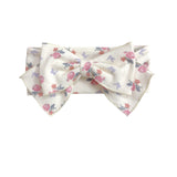 The Mia Hair Accessory by Mabel and Honey is an ivory ribbed fabric headband with a large bow, adorned with a pink and purple floral design.