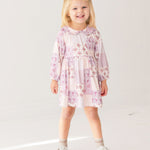 A young girl with blonde hair smiles while wearing the Liliana Floral Dress by Mabel and Honey—featuring long sleeves, a pink and white pattern, and a Peter Pan collar—paired with white socks and brown shoes against a white background.