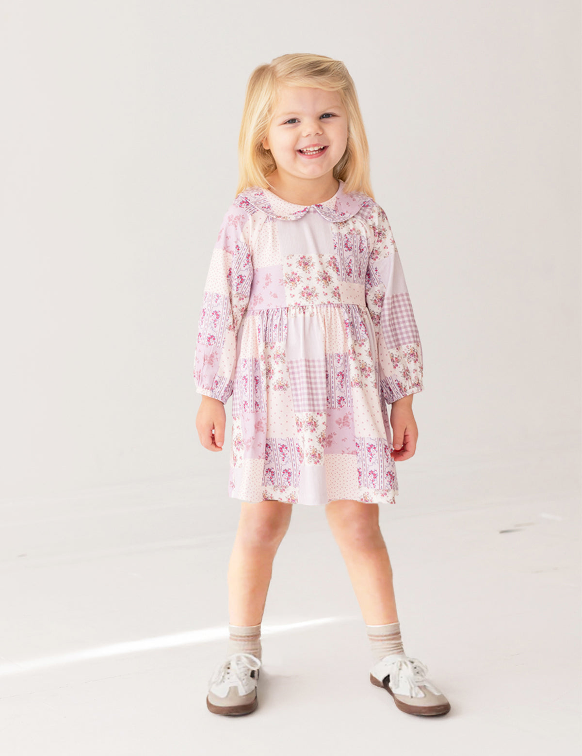 A young girl with blonde hair smiles while wearing the Liliana Floral Dress by Mabel and Honey—featuring long sleeves, a pink and white pattern, and a Peter Pan collar—paired with white socks and brown shoes against a white background.