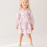 A young girl with blonde hair smiles while wearing the Liliana Floral Dress by Mabel and Honey—featuring long sleeves, a pink and white pattern, and a Peter Pan collar—paired with white socks and brown shoes against a white background.