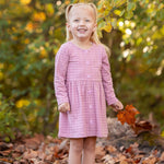 A young girl with blonde pigtails smiles among fallen autumn leaves, holding an orange leaf. She wears a Mabel and Honey Liliana Plaid Dress with green and yellow foliage in the background.