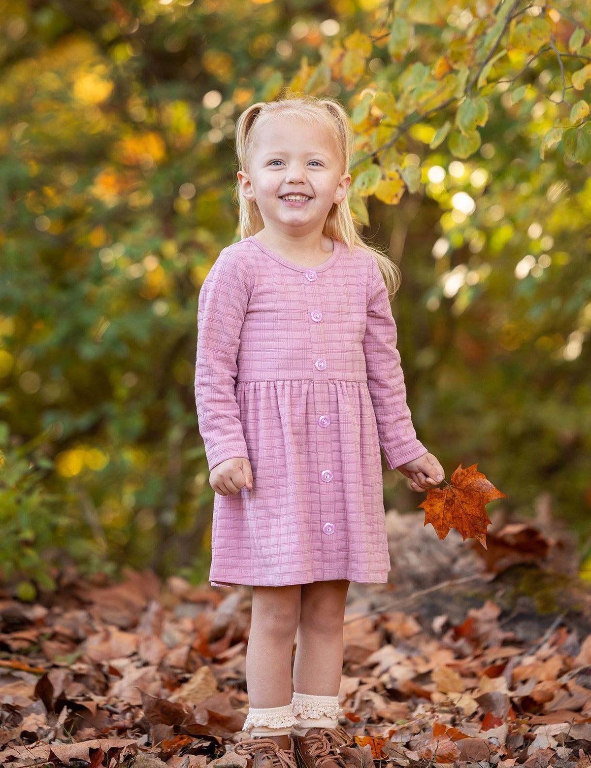 A young girl with blonde pigtails smiles among fallen autumn leaves, holding an orange leaf. She wears a Mabel and Honey Liliana Plaid Dress with green and yellow foliage in the background.
