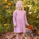 A young girl with blonde pigtails smiles among fallen autumn leaves, holding an orange leaf. She wears a Mabel and Honey Liliana Plaid Dress with green and yellow foliage in the background.