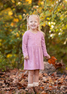 A young girl with blonde pigtails smiles among fallen autumn leaves, holding an orange leaf. She wears a Mabel and Honey Liliana Plaid Dress with green and yellow foliage in the background.
