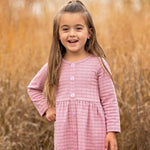 A young girl with long brown hair stands outdoors in front of tall golden grass, smiling and wearing a Liliana Plaid Dress by Mabel and Honey.