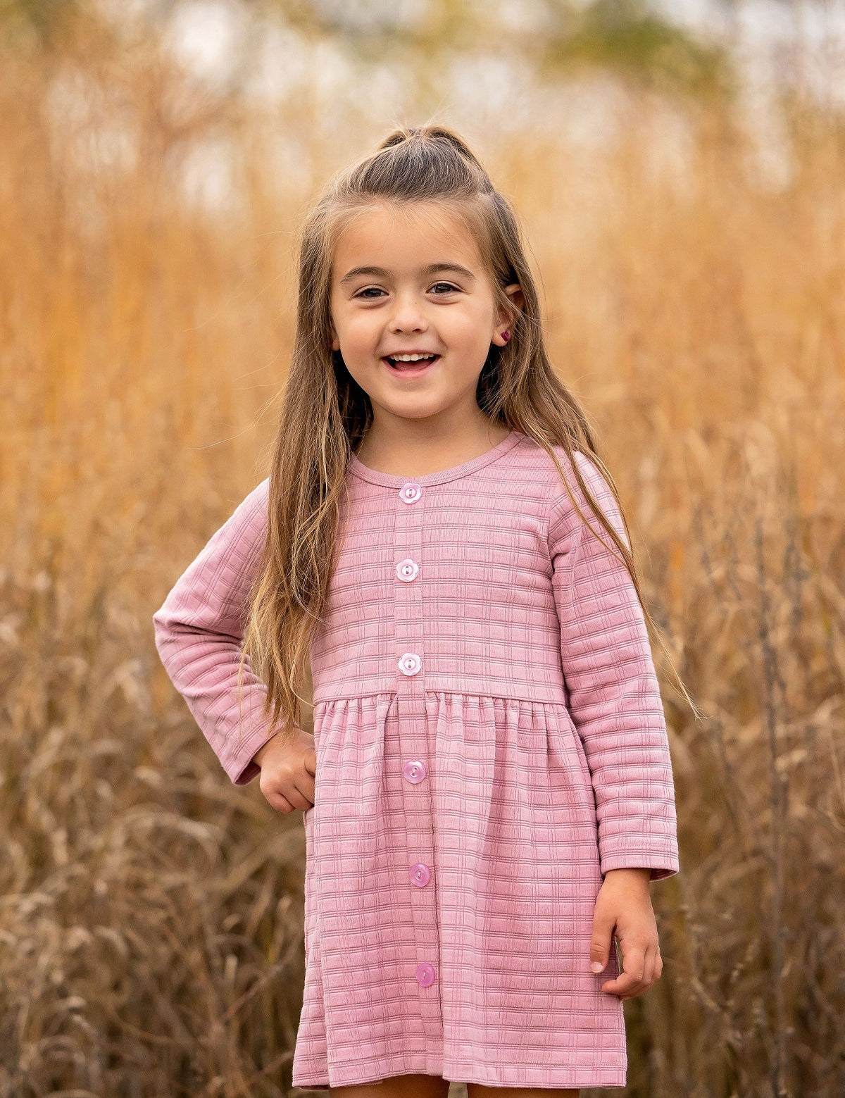 A young girl with long brown hair stands outdoors in front of tall golden grass, smiling and wearing a Liliana Plaid Dress by Mabel and Honey.