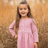 A young girl with long brown hair stands outdoors in front of tall golden grass, smiling and wearing a Liliana Plaid Dress by Mabel and Honey.