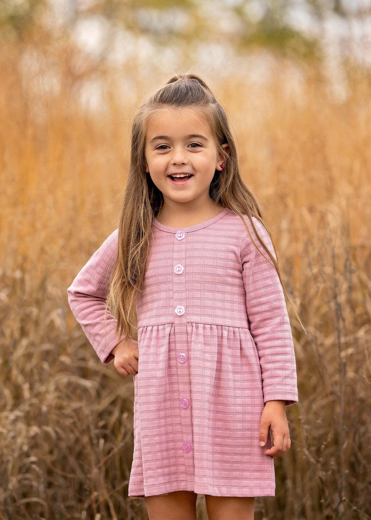 A young girl with long brown hair stands outdoors in front of tall golden grass, smiling and wearing a Liliana Plaid Dress by Mabel and Honey.
