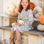 A young girl with curly red hair, wearing the Mabel and Honey Brooklyn Floral Dress and brown boots, sits on a rustic bench holding a small white pumpkin. She smiles, surrounded by white and orange pumpkins against a white wooden backdrop.