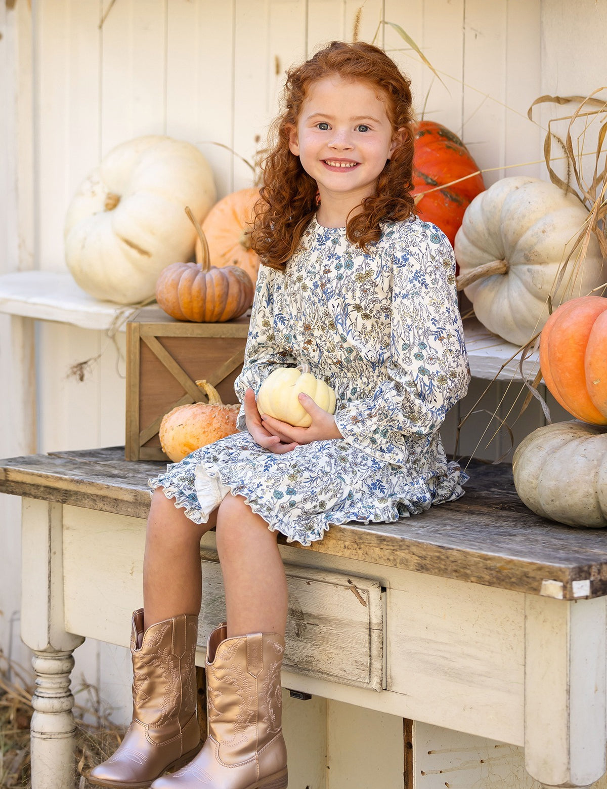 A young girl with curly red hair, wearing the Mabel and Honey Brooklyn Floral Dress and brown boots, sits on a rustic bench holding a small white pumpkin. She smiles, surrounded by white and orange pumpkins against a white wooden backdrop.