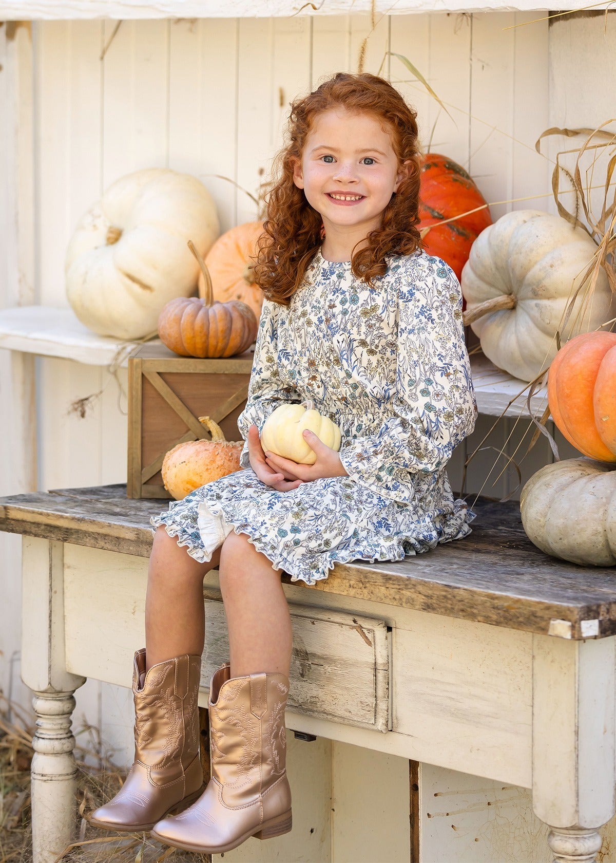A young girl with curly red hair, wearing the Mabel and Honey Brooklyn Floral Dress and brown boots, sits on a rustic bench holding a small white pumpkin. She smiles, surrounded by white and orange pumpkins against a white wooden backdrop.