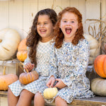 Two young girls in Mabel and Honey's Brooklyn Floral Dress sit on a bench, smiling with small pumpkins. Metallic boots and shelves of white and orange pumpkins behind them create a festive autumn scene.