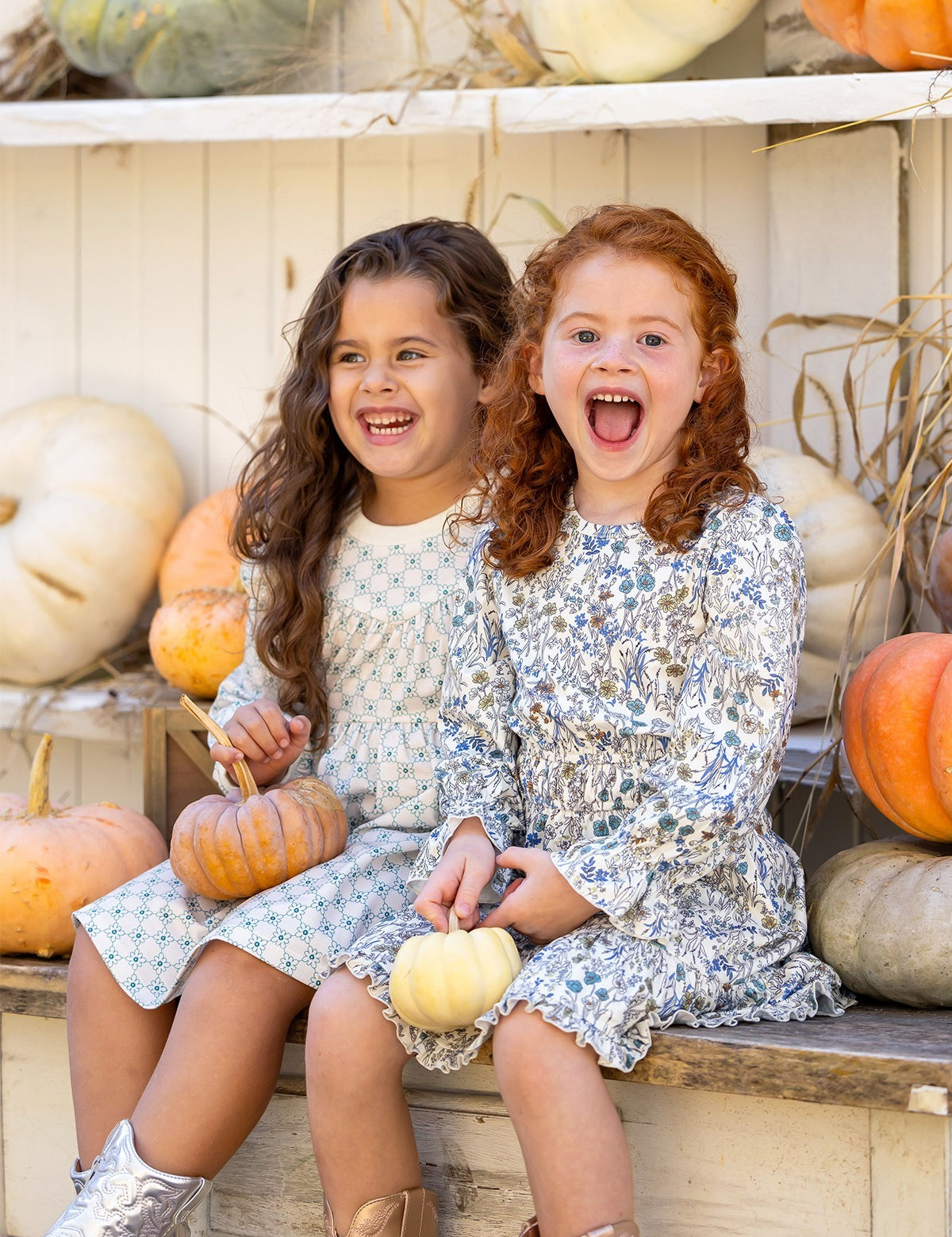 Two young girls in Mabel and Honey's Brooklyn Floral Dress sit on a bench, smiling with small pumpkins. Metallic boots and shelves of white and orange pumpkins behind them create a festive autumn scene.