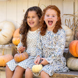 Two young girls in Mabel and Honey's Brooklyn Floral Dress sit on a bench, smiling with small pumpkins. Metallic boots and shelves of white and orange pumpkins behind them create a festive autumn scene.