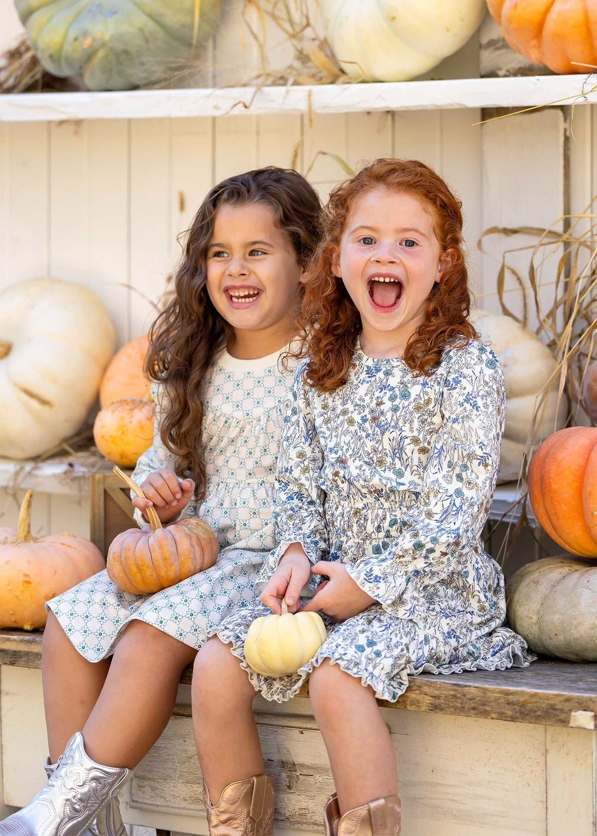 Two young girls in Mabel and Honey's Brooklyn Floral Dress sit on a bench, smiling with small pumpkins. Metallic boots and shelves of white and orange pumpkins behind them create a festive autumn scene.