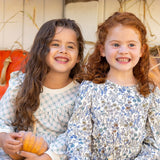 Two young girls smile brightly in their Brooklyn Floral Dresses by Mabel and Honey, each holding a small pumpkin. Orange and white pumpkins in the background add a warm autumn touch to the scene.