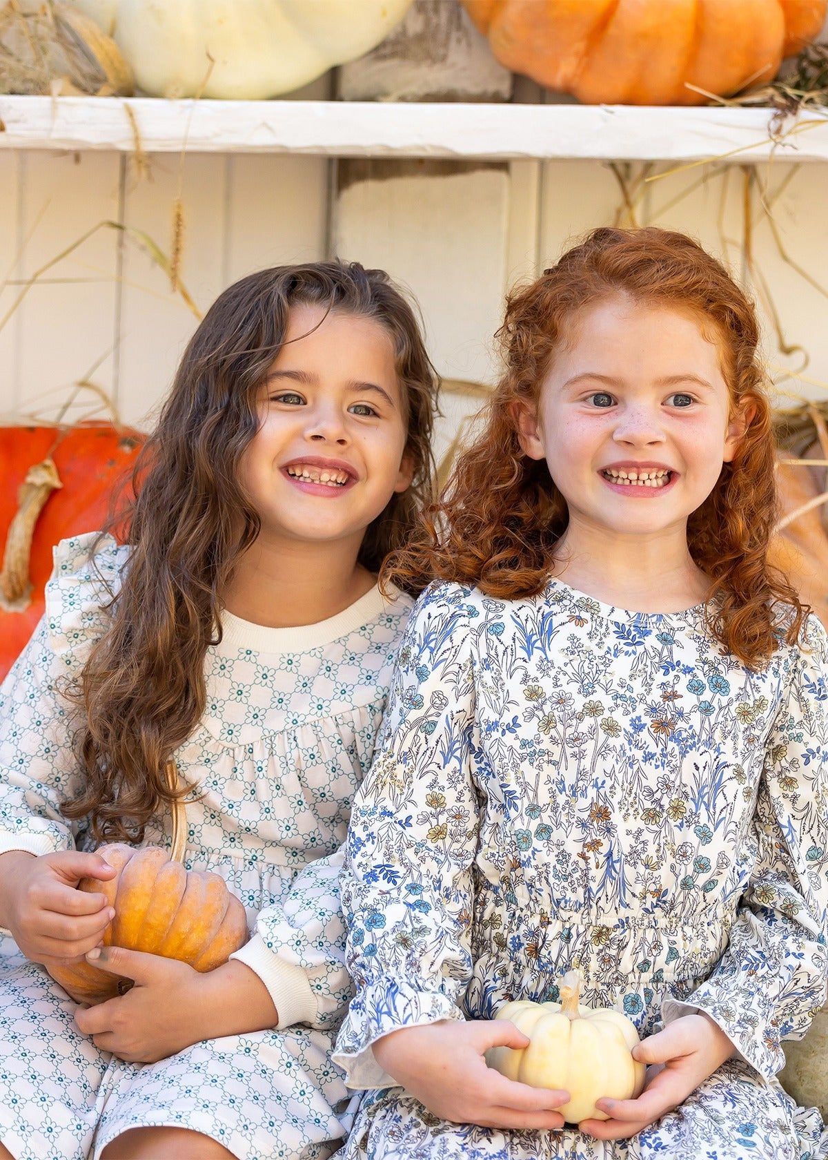 Two young girls smile brightly in their Brooklyn Floral Dresses by Mabel and Honey, each holding a small pumpkin. Orange and white pumpkins in the background add a warm autumn touch to the scene.