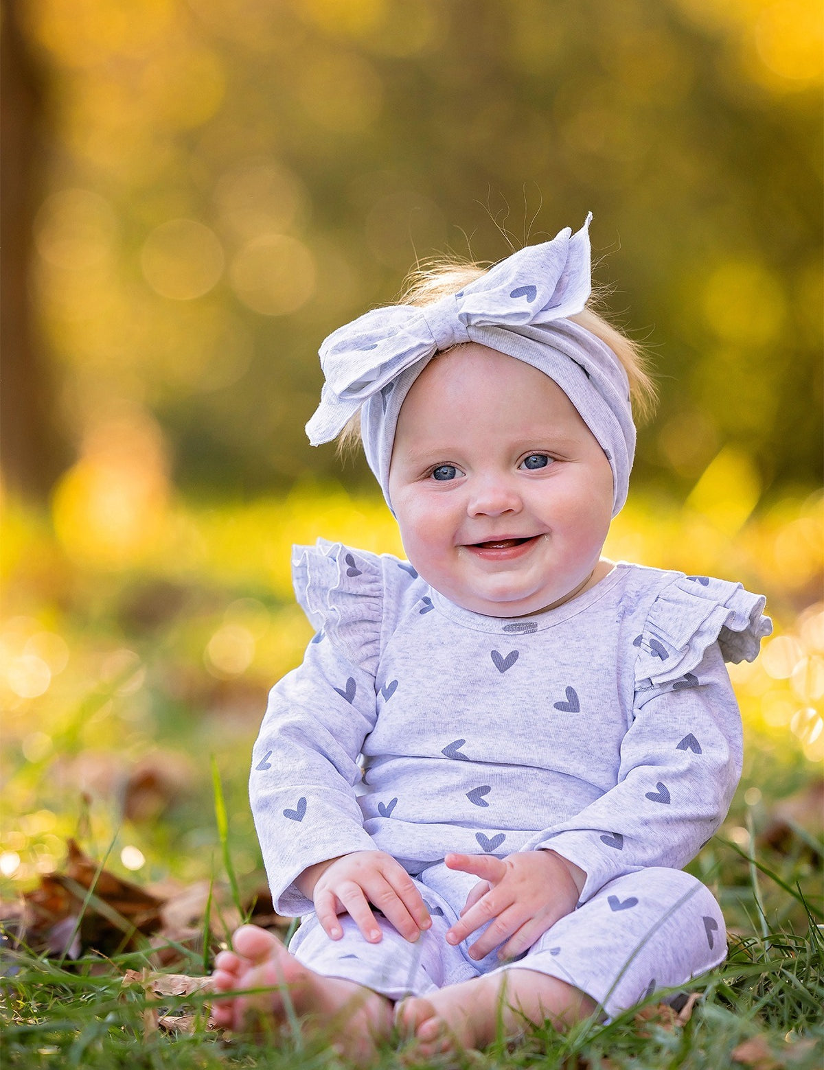 A smiling baby in the Mabel and Honey Heartfelt Romper, featuring a light gray heart pattern and ruffled shoulders with a matching headband, sits barefoot on grass against a sunlit, blurred background.