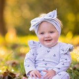 A smiling baby in the Mabel and Honey Heartfelt Romper, featuring a light gray heart pattern and ruffled shoulders with a matching headband, sits barefoot on grass against a sunlit, blurred background.