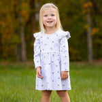 A young girl with long blonde hair stands on grass, smiling in her Mabel and Honey Heartfelt Dress, a light gray dress with small heart patterns, paired with brown shoes. Blurred trees with autumn foliage appear in the background.