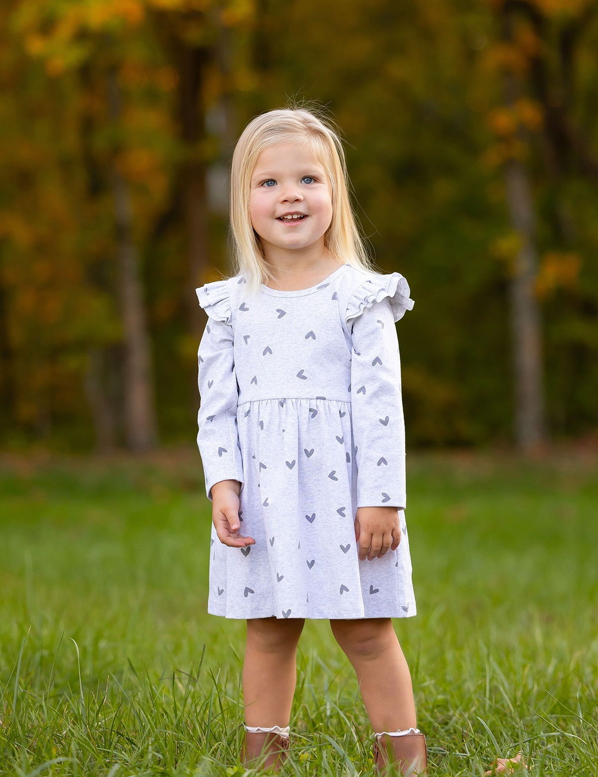 A young girl with long blonde hair stands on grass, smiling in her Mabel and Honey Heartfelt Dress, a light gray dress with small heart patterns, paired with brown shoes. Blurred trees with autumn foliage appear in the background.
