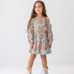 A young girl with long brown hair smiles against a white background, wearing the Mabel and Honey Riley Long Sleeve Dress with a floral print and beige lace-up boots.