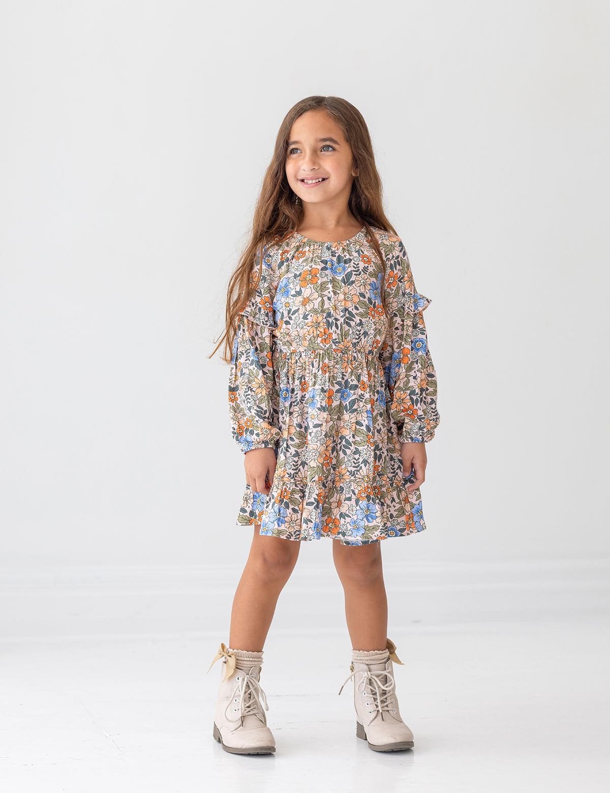 A young girl with long brown hair smiles against a white background, wearing the Mabel and Honey Riley Long Sleeve Dress with a floral print and beige lace-up boots.