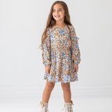 A young girl with long brown hair smiles against a white background, wearing the Mabel and Honey Riley Long Sleeve Dress with a floral print and beige lace-up boots.