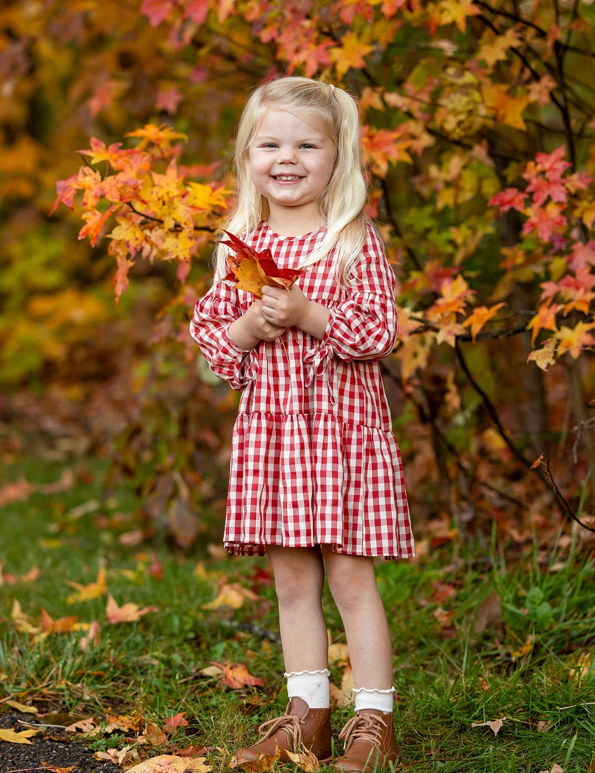 A young girl smiles outdoors, holding orange autumn leaves and wearing the Mabel and Honey Tis' the Season Dress, surrounded by colorful fall foliage.
